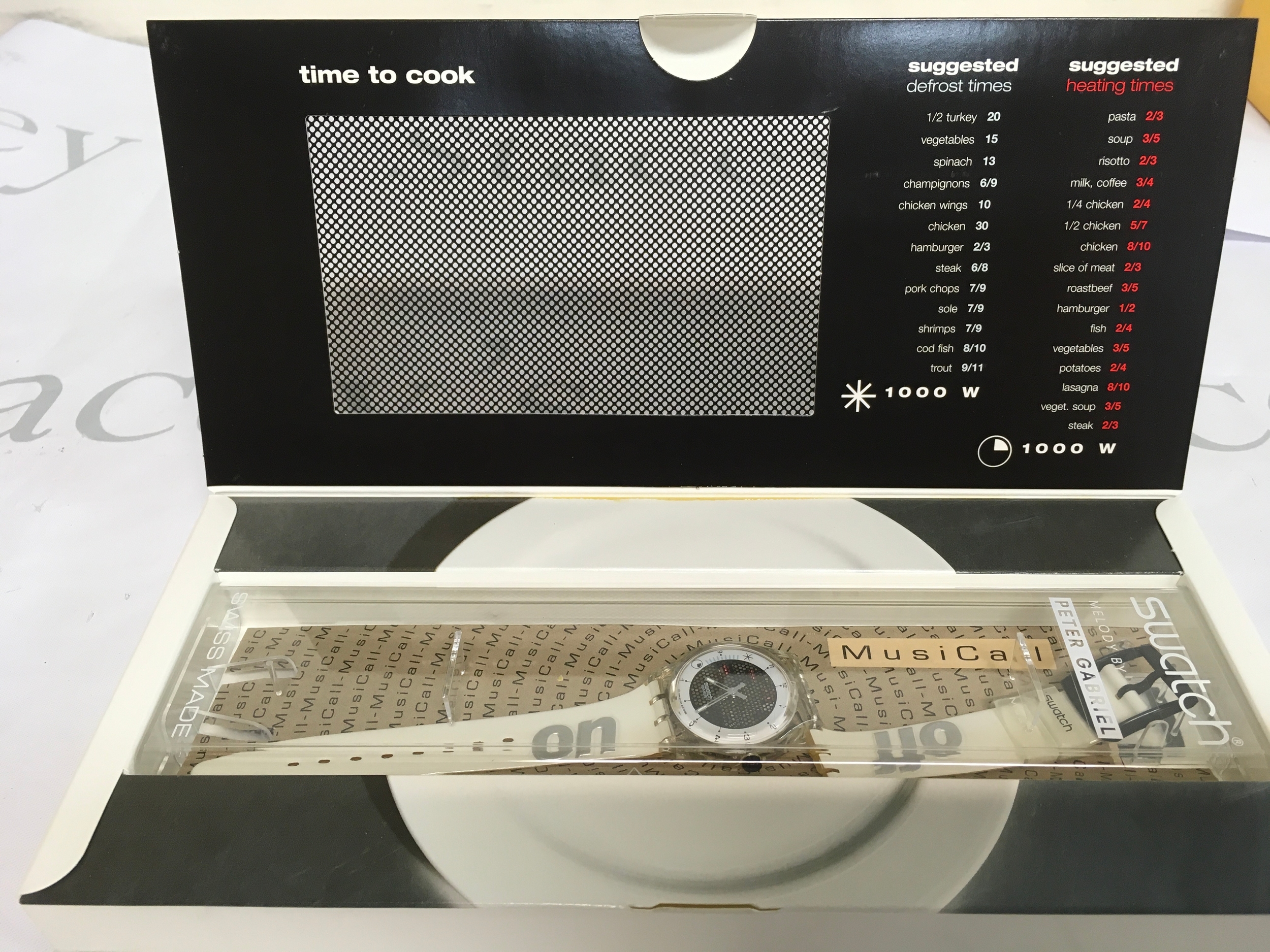 1996 Swatch Musicall watch designed by Peter Gabriel, model SLK114 "Time to Cook". The watch features a unique microwave-themed case and includes "on" and "off" text on the white strap. It comes in special packaging shaped like a microwave oven, featuring cooking times on the box flap