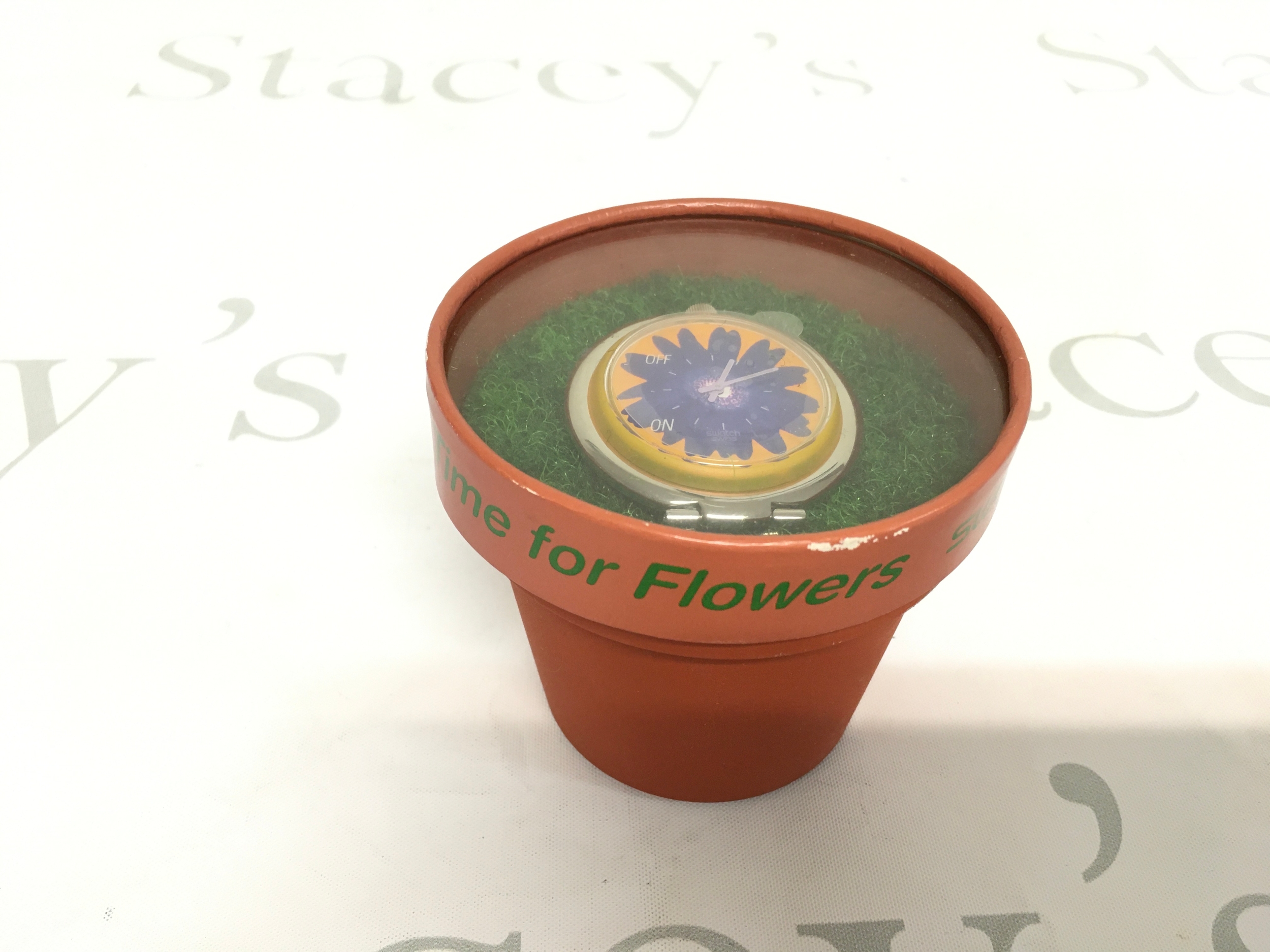 vintage 1997 Swatch "Flower Power" (PUJ101) pop table alarm clock. It features a bright blue and yellow flower design on the dial and is housed inside a terracotta-style pot.