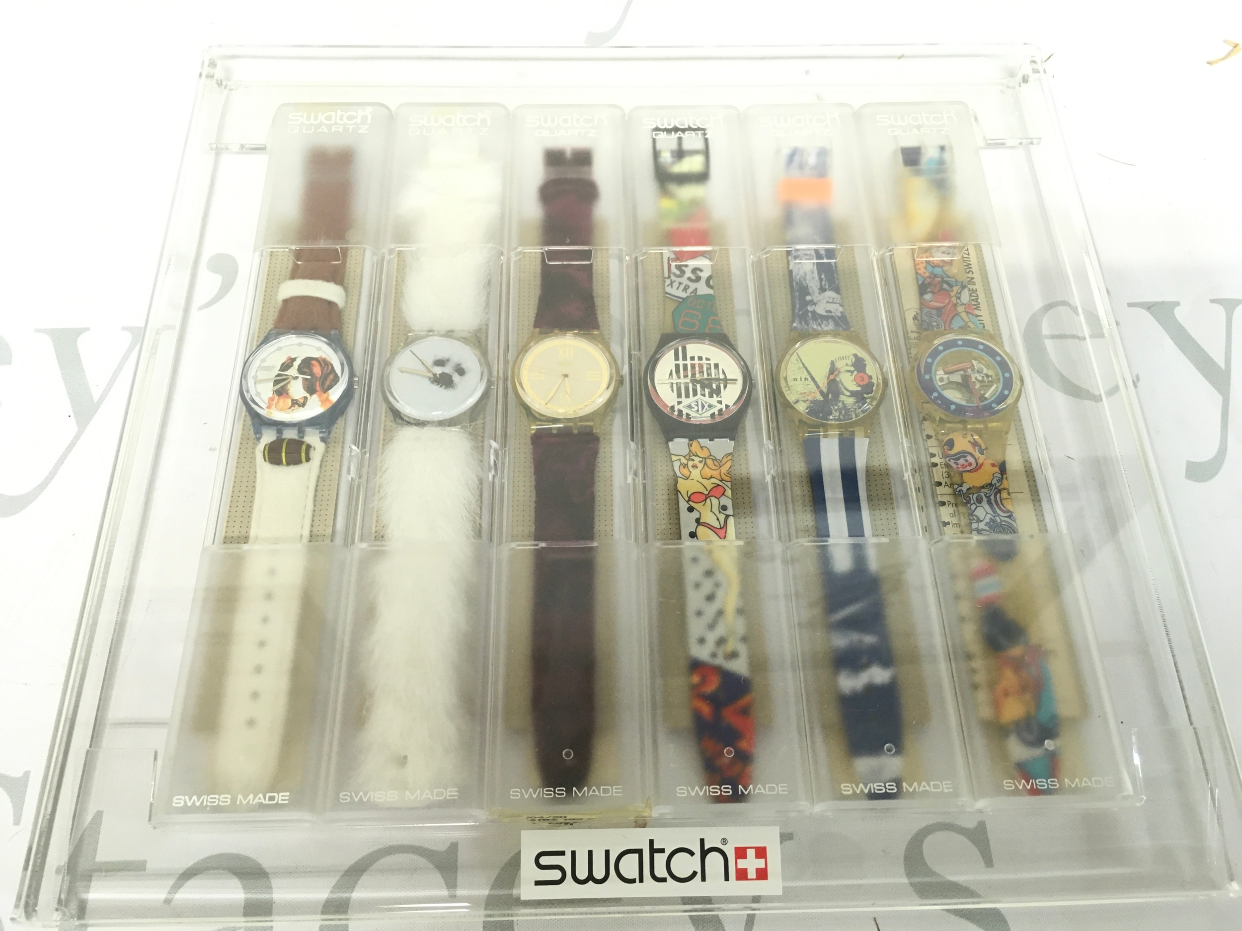 collection features six vintage Swatch Gent wristwatches from the 1990s, presented in their original transparent plastic display case. The set includes diverse designs, such as a watch with a faux fur strap resembling a sheep, a watch with a dog-themed dial, and artistic designs featuring various graphics