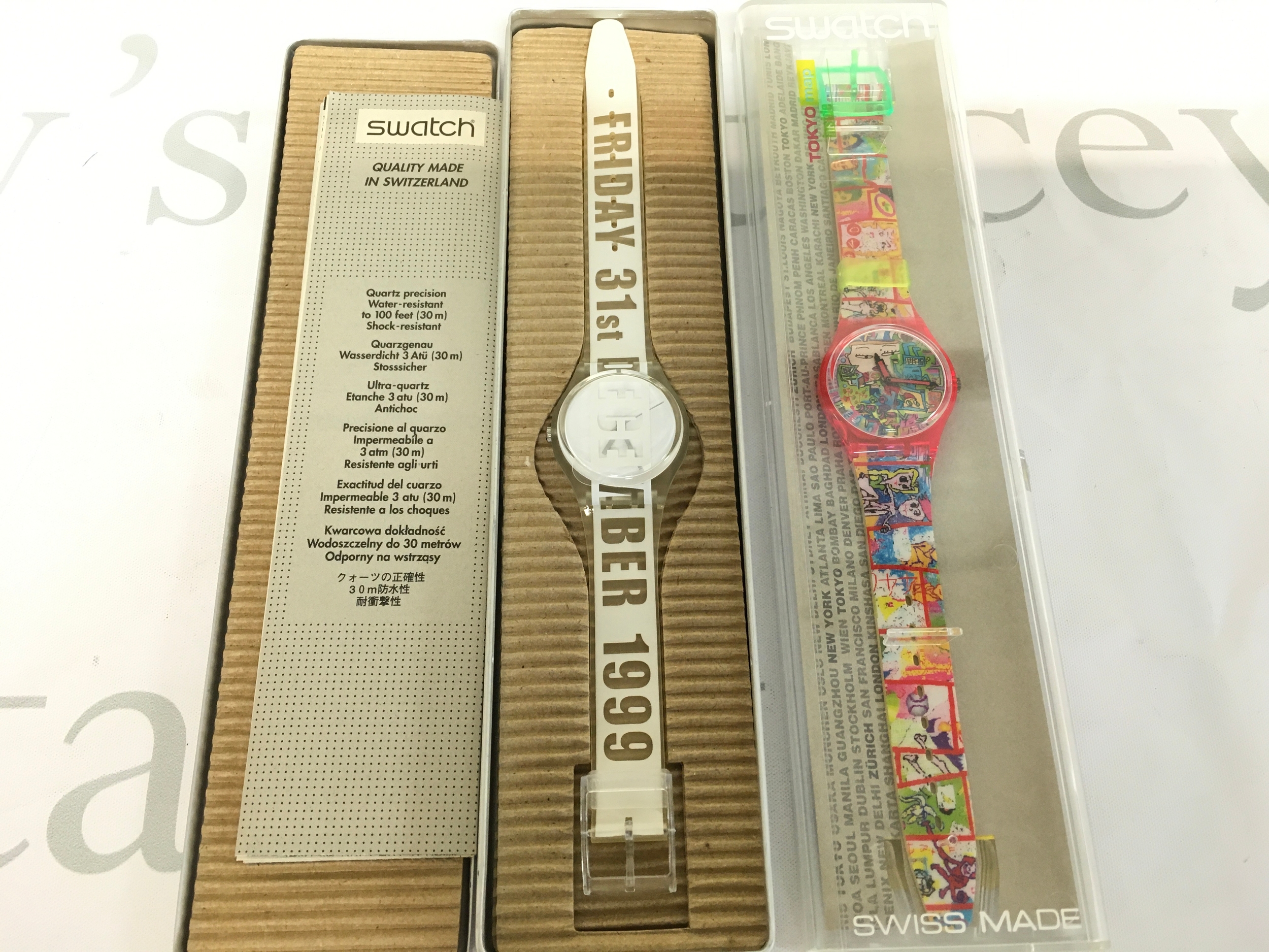 Two swatch watches including Swatch GK302 "White Card" wristwatch from 1999, featuring a transparent case and white strap printed with "FRIDAY 31st DECEMBER 1999" and 1997 Swatch Gent TOKYO MANGA GR133 watch featuring a bright, comic-book style graphic strap