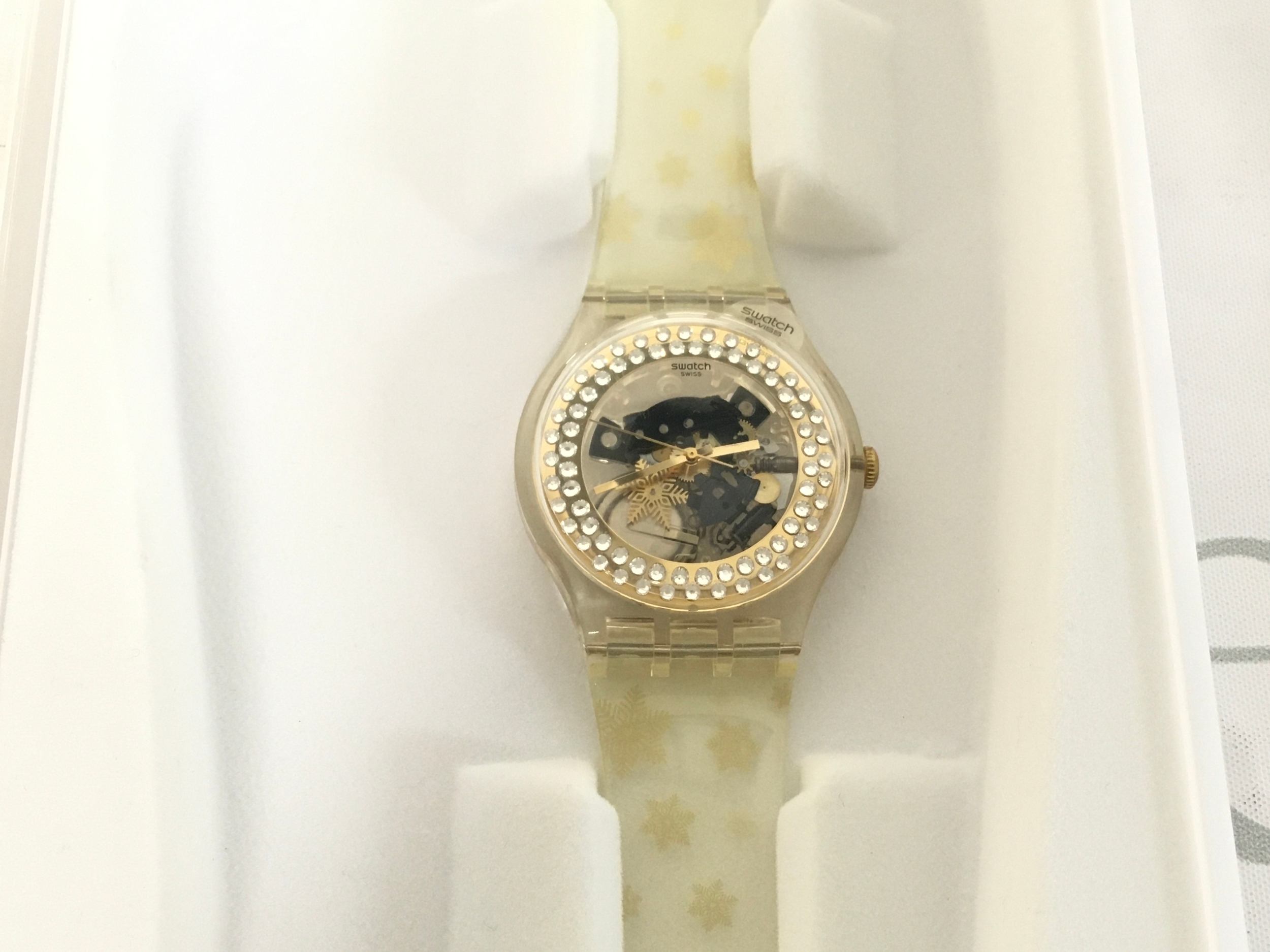 Swatch Snow Your Time Away special edition watch features a skeletonized dial with a golden rim and sparkling white crystals. It comes in unique transparent packaging, designed to look like a snowy scene. The set includes original white fleece mittens adorned with golden snowflakes. This specific Christmas special from 2010 was limited to only 9999
