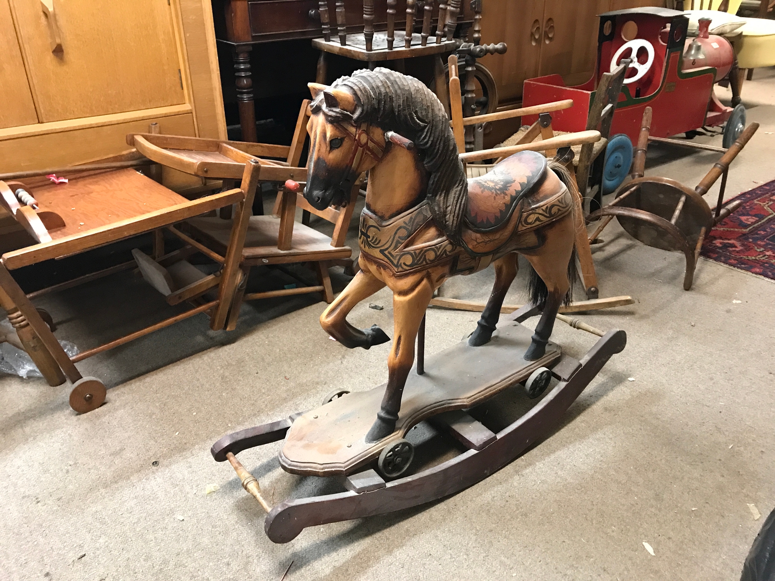 A vintage hand-carved and hand-painted wooden rocking horse featuring a decorative floral-patterned saddle, a carved mane, and an attached wooden platform with small metal wheels resting on curved rocking runners.72cm tall 84cm wide 