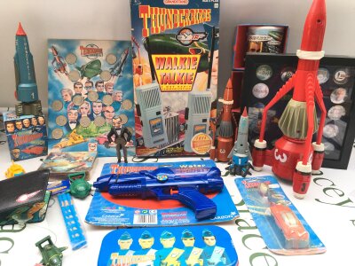 A Collection of Various Thunderbirds Toys, mugs, Stickers,. A collection Of Pin Badges In Display Case. Etc.