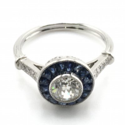 A Platinum target style ring with central old-cut diamond surrounded by calibre cut sapphires in a beaded setting, with further diamonds to the shoulders. Centre diamond 0.56ct. Sapphires 0.60ct. Remaining diamonds 0.07ct. Total weight 4.6g, size M. Postage category A