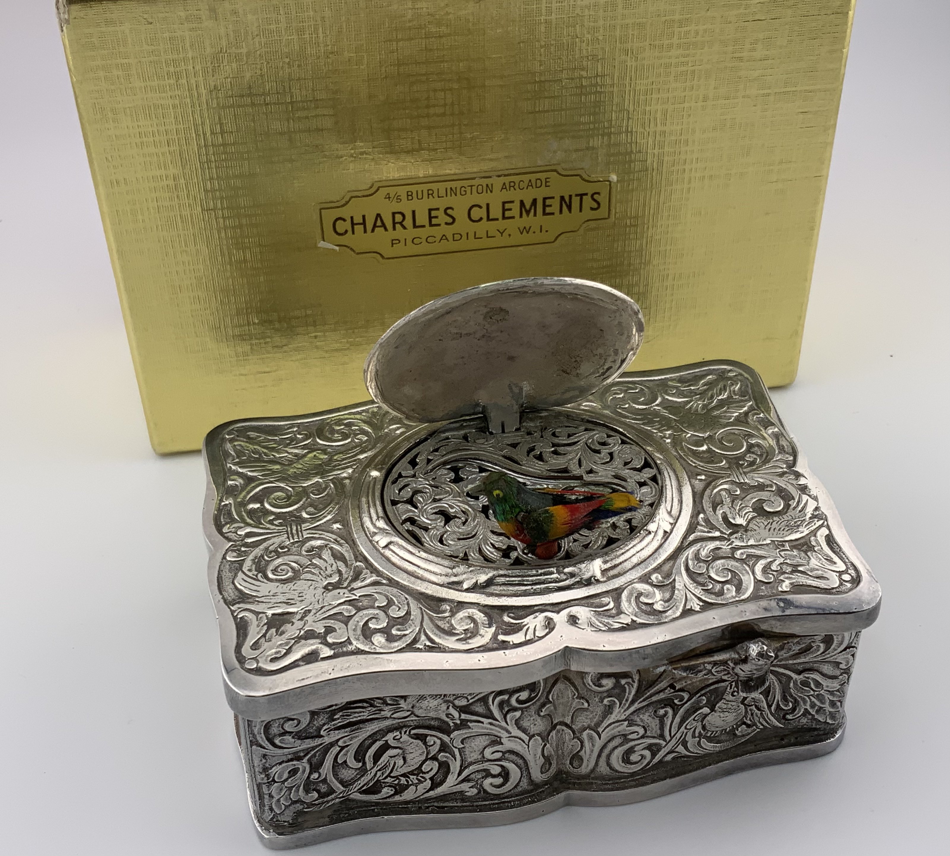 An ornate Swiss white metal ''Singing Bird'' automaton music box, stamped ''METAL'' and ''EB'' in an oval, rectangular, profusely embossed with foliate and exotic bird designs, with instruments and cherubs, the oval hinged cover opening to reveal an automated and feathered singing bird not in working order.