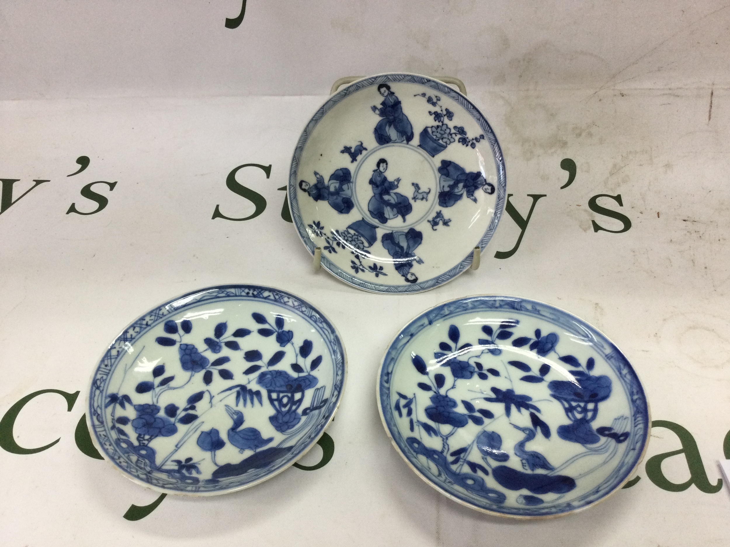 Three Kang Xi saucers  one depicting women with dogs and potted flowers within a blue border verso with leaf decoration and four character mark to centre within concentric circles, 13.5Cm chip and hairline crack to the rim. the other two depict a stork amongst floral and symbolic objects,both with minor flitting to the rim 13cm.