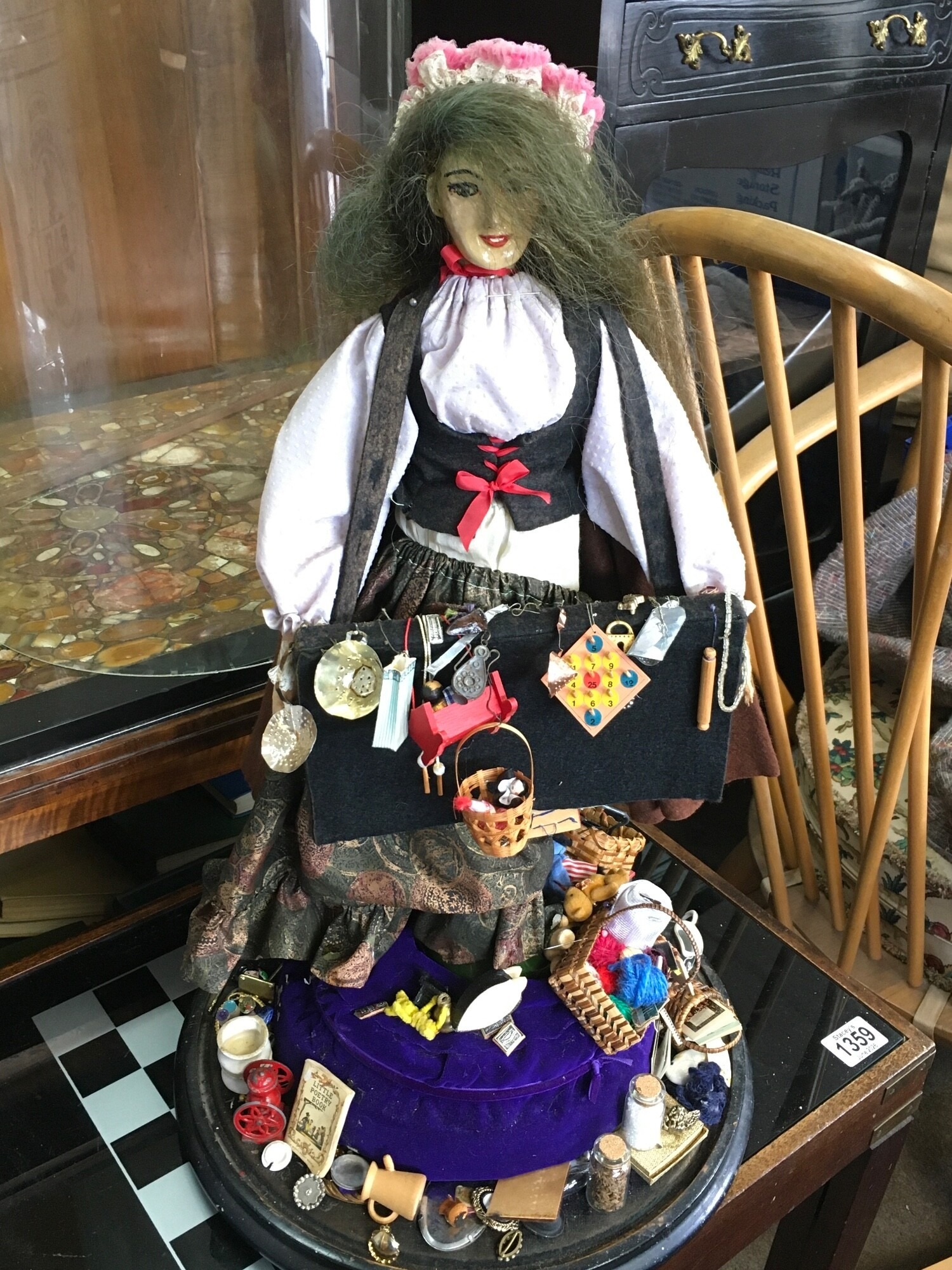 A Pedlar doll figure in a glass display dome, with an assortment of miniature trinkets. Dome is 67cm tall