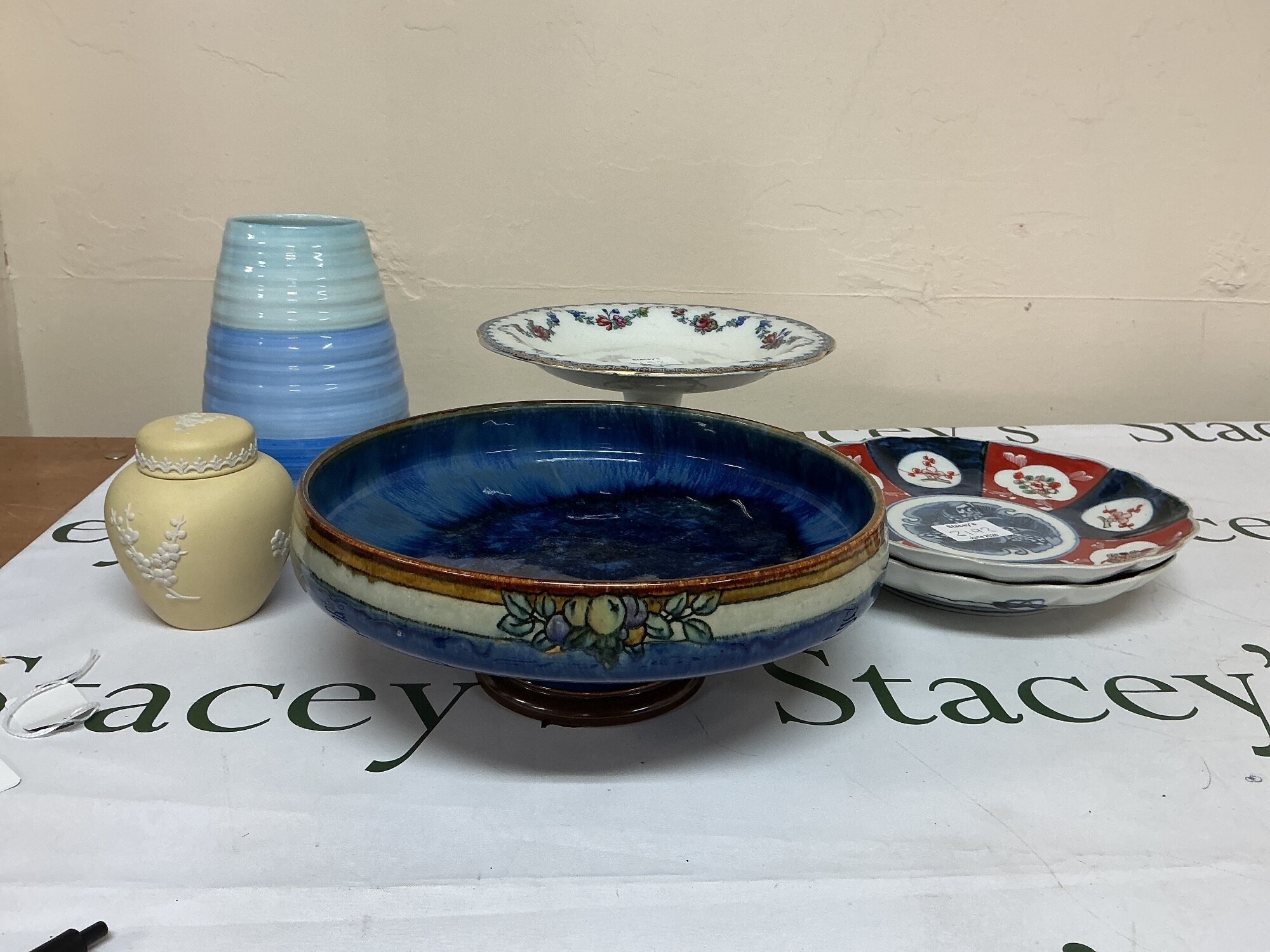 A collection of a mixed lot of ceramic decorative items featuring Imari Style porcelain plates, wedge wood primrose jasperware ginger jar, pedestal cake stand, a modern banded studio pottery vase, and a large Art Deco style ceramic fruit bowl.