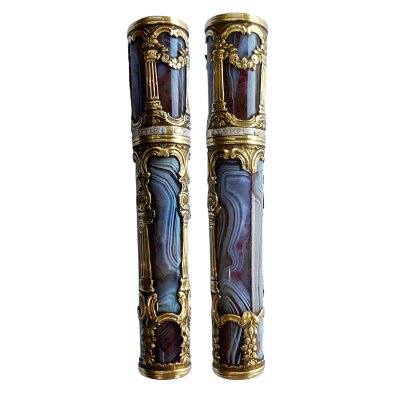 An agate needle case with enamelled gold mounts, English, circa 1760, of cylindrical tapering form, the grey figured agate body decorated with chased borders of alternating c-scrolls and flowers and Corinthian columns around top and bottom of base and lid, the latter with a fine white enamel band with gold inscription reading: Vous Loffre L’amitite (You Offer Freindship), slight domed top and underside.
10.6cm. Some damage to agate panels and gold work.