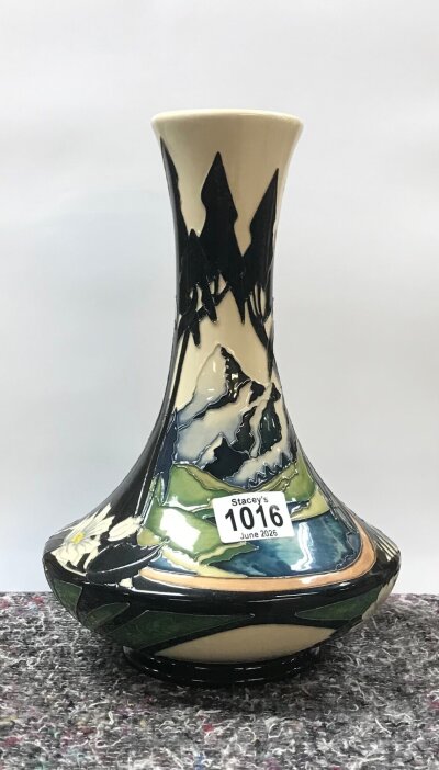 A Moorcroft vase with deep glossy colours. The pattern shows tall black silhouetted tress, a water scene with hills/ mountains and white flowers around the base. (No obvious damage or restoration)