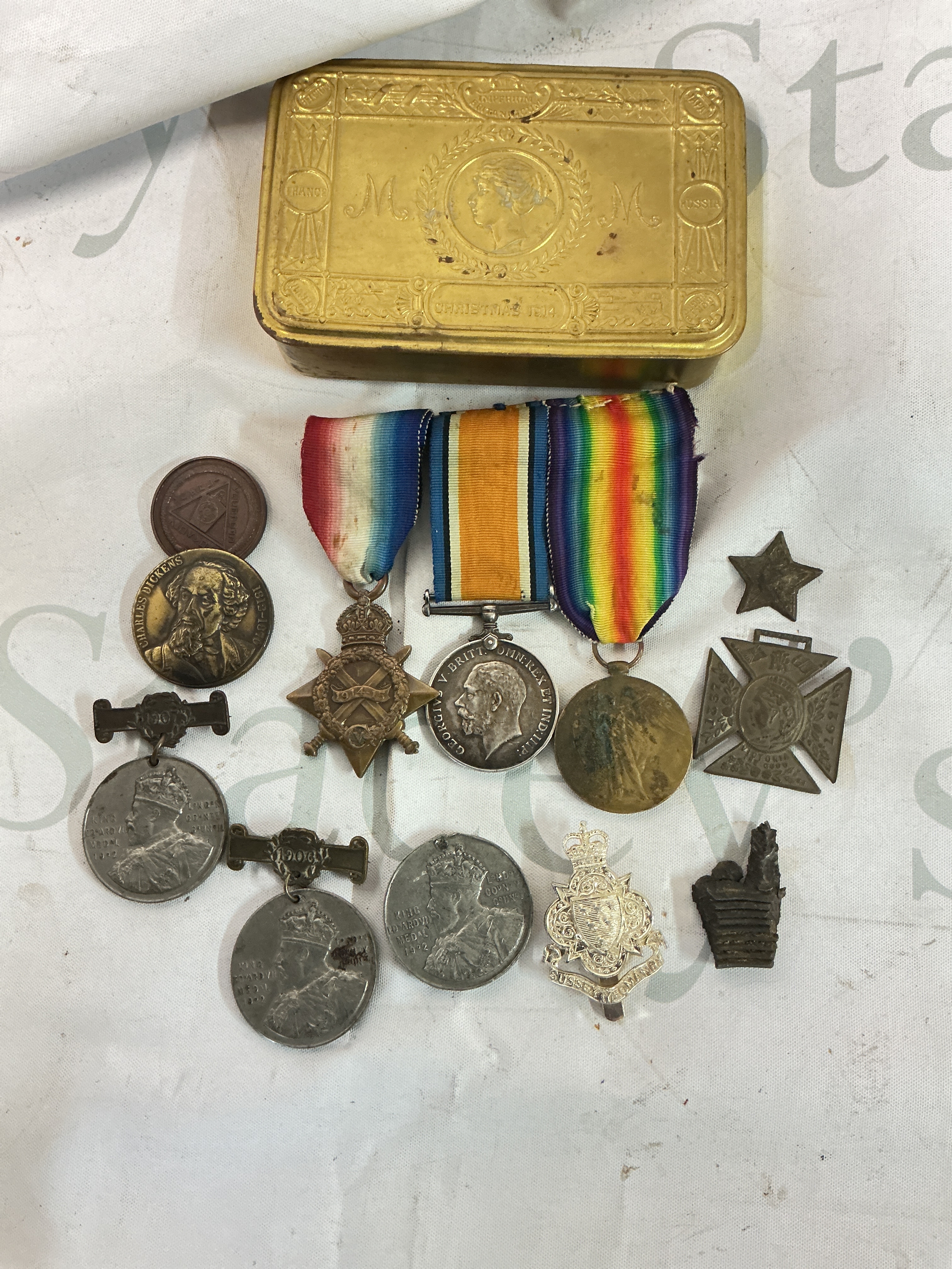A Mary Tin Containing WW1 Medal Bar, Cap Badge, Piece Of Shrapnel, 3 Coronation Medals, Charles Dickens Medallion And Queen Victoria Medallion And Two Others.