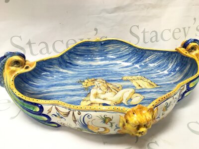An Italian maiolica centrepiece bowl, hand-painted classical scene in the interior and is adorned with mythical fish-shaped handles. 46cm wide 13cm tall