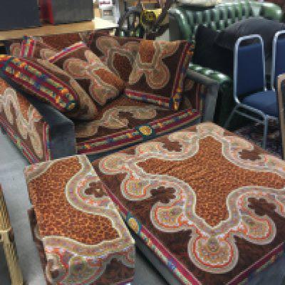 A three piece Versace suite including two sofas, a large chair and ottoman upholstered in a orange, brown , red, green and blue Gianni Versace velvet fabric featuring a leopard and Medusa print. Five cushions and two side fitting coffee tables. Also includes five non matching cushions. Dimensions - Sofas 228cm wide 70cm tall 125cm long , other sofa 225cm wide 60cm tall 118cm deep. Chair 130cm wide 75cm tall, 108cm long. Ottoman 102cm wide 36cm tall 100cm long. Side tables 65cm wide 26cm tall.