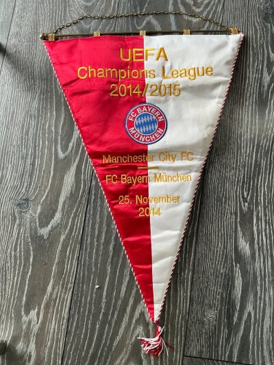 2014 - 2015 Manchester City v Bayern Munich Exchange Football Pennant: Large attractive pennant given by Phillip Lahm to Vincent Kompany immediately before kick off in the traditional pre match exchange. Match details date and Champions League 2014/2015 embroidered either side of Bayern Munich cloth badge.