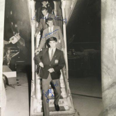 George Best Signed Manchester United Press Photo: Best Foulkes and Cantwell are coming down the stairs of the airplane in August 1964 in Hamburg. All 3 have clearly hand signed without dedication. Press stamp and German annotations to rear.