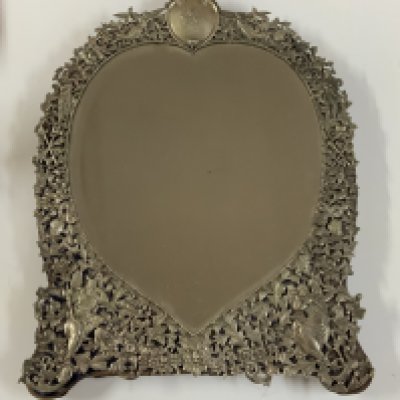 An Antique Victorian hallmarked Sterling Silver dressing table mirror having hand-chased angels, God&#2013266057۪s and Goddesses, scroll-work, and floral decoration, with a top engraved cartouche, an original bevelled heart-shaped mirror on a blue velvet backing with easel stand. Made by William Comyns of London in 1887. Height. 51.5cm x 37cm.
