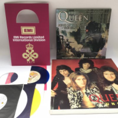 A box of Queen related items comprising coloured vinyl, books, clock etc plus other coloured 7" singles and a Phil Collins CD/VHS box set.