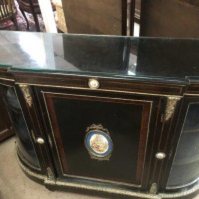 A Victorian ebonised credenza with two glass doors on either side with shelves for storage, gold ormolu trim and plaques ,porcelain hand painted panels of floral imagery and in the middle a classical figures fishing scene ,with a glass top. A foot is loose and needs reattaching. 150cm wide 98cm tall.